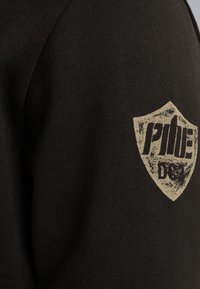 Sudadera negra con un tejido texturizado. Presenta un logo en forma de escudo dorado en la manga con texto estilizado. Diseño simple y casual.