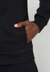 Sudadera negra con bolsillo canguro, puños y dobladillo acanalados, hecha de un suave tejido texturizado, con un detalle en la costura lateral.