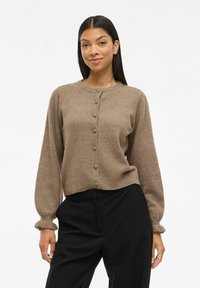 Femme portant un cardigan marron à boutons avec des manches bouffantes et un pantalon noir taille haute, se tenant devant un fond blanc uni.