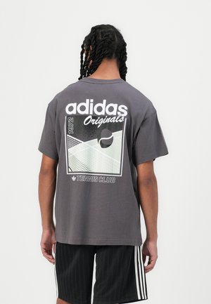 Osoba z warkoczami ubrana w ciemnoszarą koszulkę Adidas Originals z grafiką kortu tenisowego i piłki na plecach.