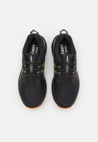Scarpe da corsa nere con tomaia in rete, accenti verdi e dettagli arancioni. Presentano un design con lacci e una suola in gomma testurizzata.