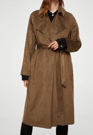 Kvinde iført en lang brun trenchcoat med bælte over en sort skjorte og sorte bukser, står mod en ensfarvet baggrund.