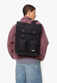 Svart ryggsäck tillverkad av hållbar textil, med två framfickor, justerbara remmar och ett vikbart lock med tryckknappar.