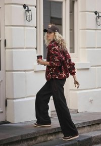 Maroon faux fur jacket with geometric pattern, black wide-leg pants, and brown sneakers, holding a coffee cup, walking near a building.