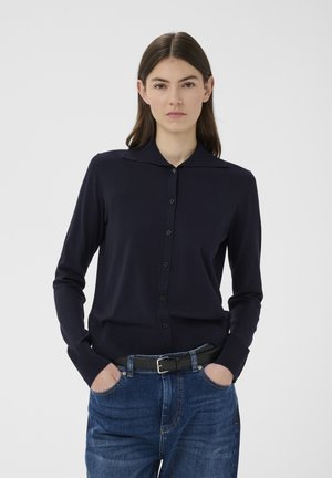 Nő egyenes barna hajjal, sötétkék, gombos kardigánban és kék farmernadrágban fekete övvel, kezét a zsebében tartja, semleges háttér előtt.