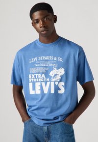 Tricou din bumbac albastru cu text și imagini grafice albe, având inscripțiile "Levi Strauss & Co." și "Extra Strength Levi's." Croială lejeră, mâneci scurte.