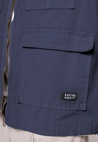 Chaqueta azul marino con tejido texturizado, cierre de cremallera y dos bolsillos frontales. La etiqueta en un lado dice "KAOTIKO SOCIETY."