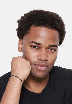 Schwarzes geflochtenes Armband am Handgelenk mit Kettendesign. Das Model hat kurzes, lockiges Haar und trägt ein schwarzes T-Shirt vor einem weißen Hintergrund.