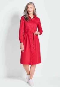 Rotes Hemdkleid mit langen Ärmeln, tailliertem Gürtel und knielanger Saumlänge, aus glattem Material, kombiniert mit weißen Sneakers.