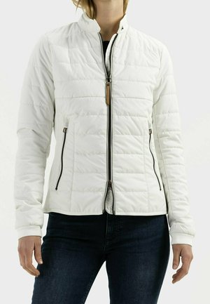 Personne portant une veste matelassée blanche avec une fermeture éclair à l'avant et des poches latérales zippées, associée à un jean bleu foncé.