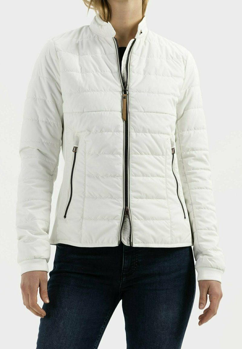 Personne portant une veste matelassée blanche avec une fermeture éclair à l'avant et des poches latérales zippées, associée à un jean bleu foncé.