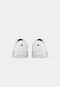 Ett par vita låga sneakers med logotyp på hälen och präglat varumärkesnamn på sulorna, visade från baksidan på vit bakgrund.