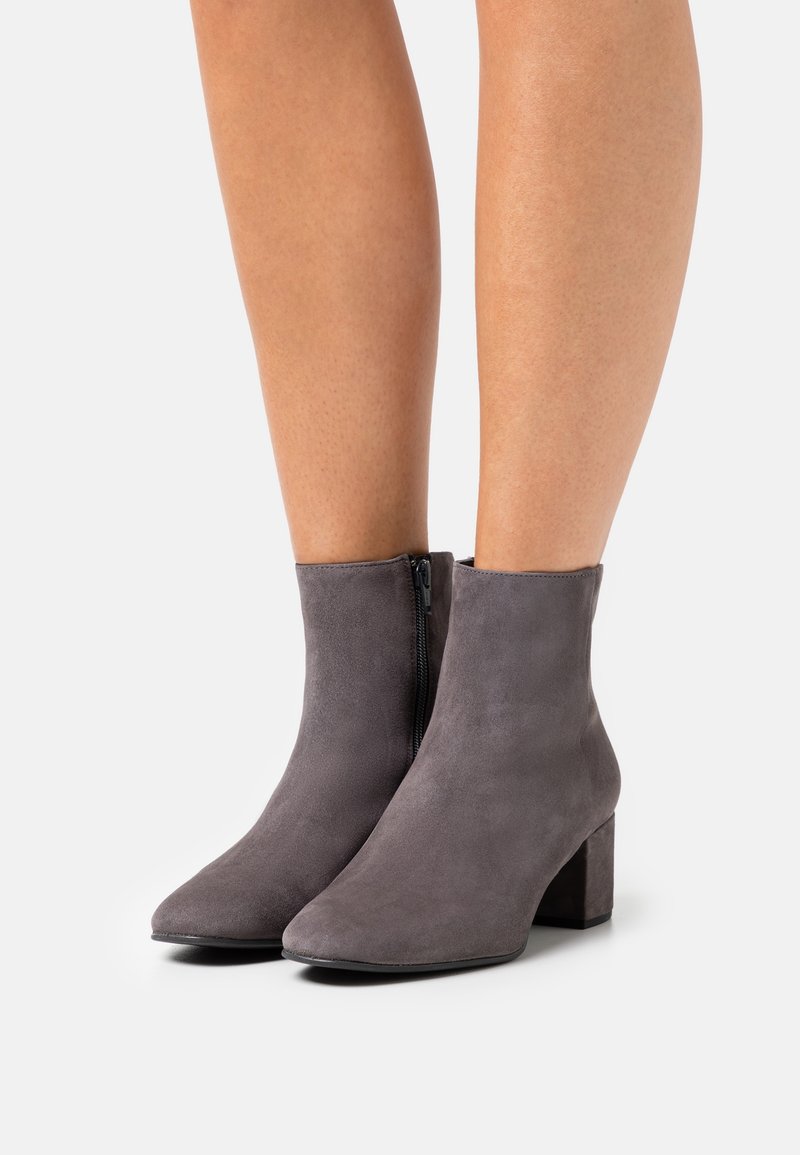 Bottes grises en daim montant à la cheville avec talons carrés et fermetures éclair latérales, portées sur des jambes nues sur un fond blanc uni.