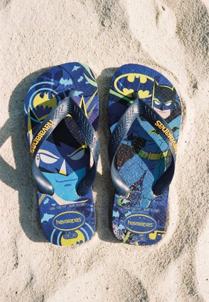 Dziecięce niebieskie japonki Havaianas z motywem Batmana, leżące na jasnym piasku z grafikami kreskówkowymi i żółtymi logo.