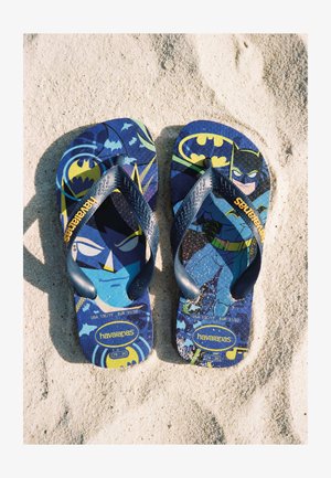 Tongs bleues pour enfants à thème Batman de Havaianas reposant sur un sable clair avec des graphiques dessin animé et des logos jaunes.