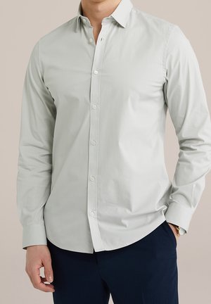 Homme portant une chemise boutonnée à manches longues gris clair et un pantalon foncé, une main dans la poche, sur un fond neutre.