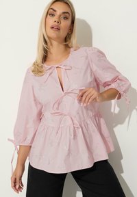 Blusa rosa realizzata in un tessuto leggero, con fiocchi argentati ricamati su tutta la superficie, con nodo al collo e vita e maniche arricciate.