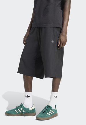 Zwarte sportbroekjes van lichtgewicht stof, met een subtiele Adidas-logo, gecombineerd met groene suède sneakers en witte sokken.