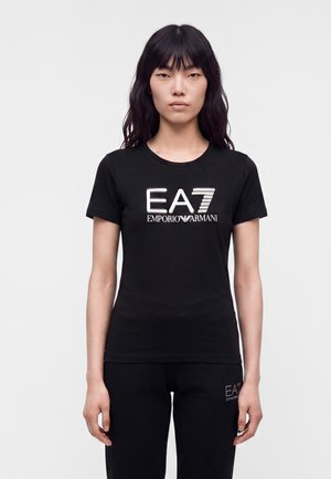 Kvinde med langt sort hår har en sort t-shirt med korte ærmer og bukser med sølvfarvede EA7 Emporio Armani-logoer foran.