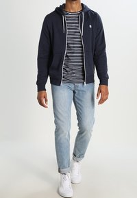 Felpa con zip color navy e cordini bianchi, abbinata a una maglietta a righe, jeans blu chiaro e sneakers bianche. Tessuto morbido, vestibilità casual.
