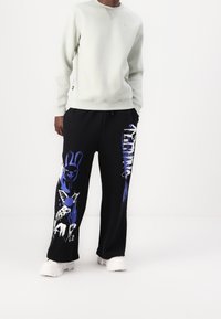 Ljusgrå sweatshirt med ribbade mudd, tillsammans med svarta träningsbyxor med blå abstrakta grafiker och en lös passform. Vita sneakers.