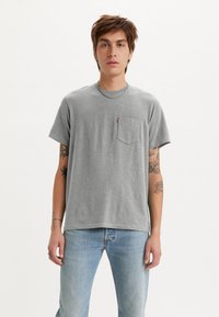Grå bomulds t-shirt med korte ærmer, rund halsudskæring, frontlomme og diskret branding. Bruges med lyseblå denim jeans.