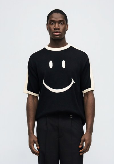 Νέος άνδρας που φοράει μαύρο πλεκτό μπλουζάκι με κοντά μανίκια και μεγάλο λευκό γραφικό με χαμογελαστή φατσούλα, καθώς και μαύρο παντελόνι που ταιριάζει, στέκεται μπροστά σε απλό φόντο.