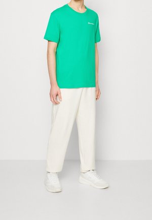Man draagt een felgroen Champion T-shirt met korte mouwen, witte losse broek en witte sneakers, staand tegen een effen lichte achtergrond.