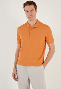 Oranžové polo tričko s klasickým límečkem, krátkými rukávy, tříknoflíkovou légou a hladkým povrchem, ve spojení s světlými kalhotami.