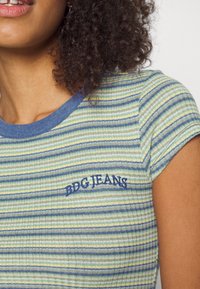 Pruhovaný top s krátkými rukávy v zelené, modré a béžové barvě s žebrovanou texturou. Na hrudi vyšitý nápis "BDG JEANS" v námořnicky modré nitce.
