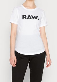 Bílá krátká rukávová tričko vyrobené z bavlny, s velkým černým grafickým prvkem "RAW." přes hruď, kulatým výstřihem a zaobleným lemem.