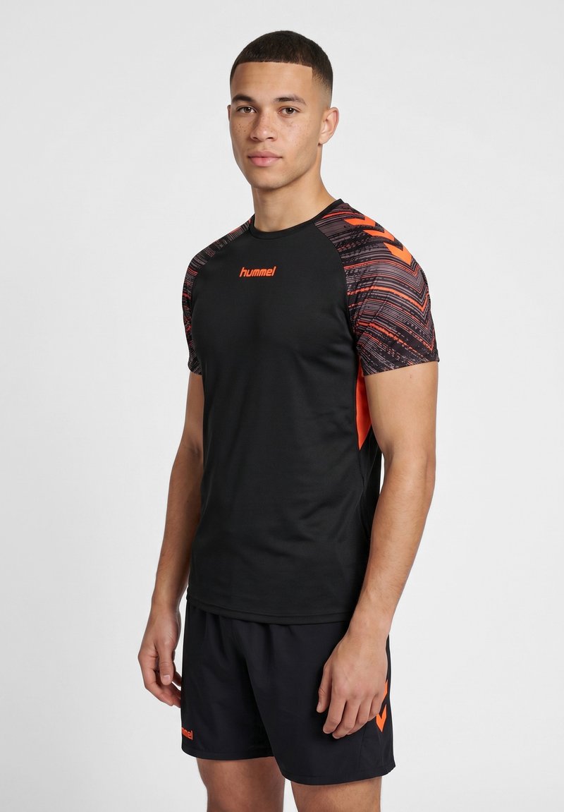 Ung mand iført sort sportstøj med orange mønstrede ærmer og logo, stående mod en ensfarvet grå baggrund.