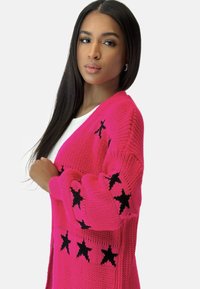 Cardigan en tricot rose avec des motifs d'étoiles noires. Présente un devant ouvert, des manches longues et des poignets côtelés pour une texture additionnelle.