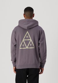 Sudadera con capucha gris que presenta un gran logotipo triangular con "H", "U" y "F" en color crema. Tejido suave con puños y dobladillo acanalados.