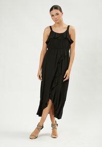 Maxi dress nera con spalline regolabili, scollatura arricciata e orlo asimmetrico; indossato con sandali con cinturini. Tessuto liscio.