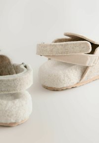 White fleece slippers with beige Velcro straps and a textured surface. Features a soft upper and a rubber sole for grip.