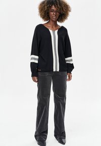 Zwarte gebreide trui met witte en grijze verticale strepen, brede mouwen en een V-hals. Gecombineerd met donkergrijze ribfluwelen broek en zwarte laarzen.