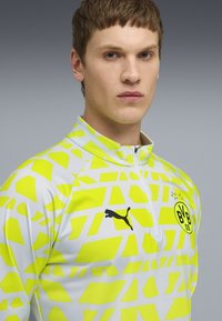 Långärmad sporttröja med ett gult geometriskt mönster på en vit bas, med hög krage, dragkedja och svart Puma-logotyp.