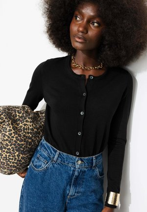 Femme avec coiffure afro portant un cardigan noir boutonné, un jean bleu, un collier chaîne en or et tenant un sac imprimé léopard.