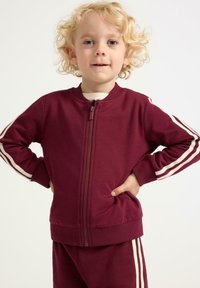 Ungt barn med krøllet blond hår iført en maroon træningsdragt med hvide striber, stående med hænderne på hofterne foran en ensfarvet baggrund.