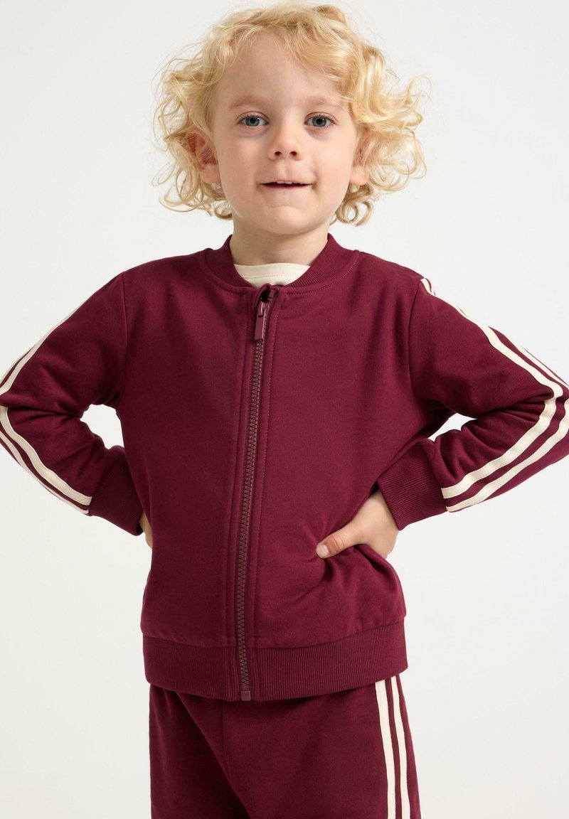 Ungt barn med krøllet blond hår iført en maroon træningsdragt med hvide striber, stående med hænderne på hofterne foran en ensfarvet baggrund.