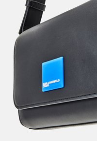 Zwarte schoudertas van glad materiaal met een vierkant blauw logo label met de tekst "KARL LAGERFELD JEANS". Rechthoekige vorm met gestikte accenten.