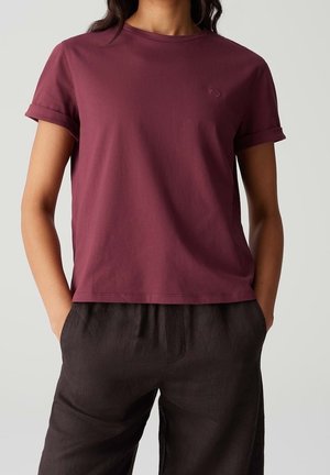 Kvinde iført en ensfarvet mørkelilla t-shirt med korte ærmer og en lille hjertebroderi på brystet samt mørkebrune bukser med hænderne i lommerne.