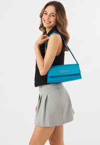 Turkoosi krokotiilikuosis laukku, jossa on läppäsuljenta ja säädettävä olkahihna. Etupuolella on erottuva logo.