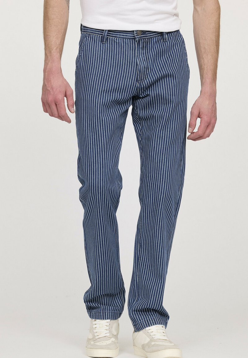 Un pantalon rayé marine présente une coupe slim, de légères rayures horizontales, des passants de ceinture et une fermeture à bouton à l'avant. Porté avec des baskets blanches.