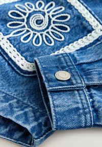 Denim jacket cuff with metal snap button, featuring white embroidered floral and geometric patterns on blue fabric.