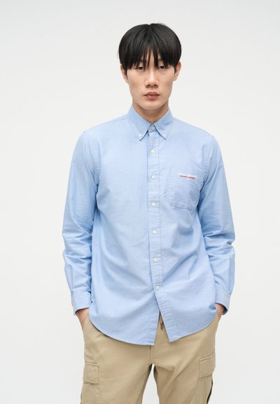 Άνδρας που φοράει ανοιχτό μπλε πουκάμισο με κουμπιά και λογότυπο "Polo Sport Ralph Lauren" και μπεζ παντελόνι καμπαρντίνας, στέκεται με τα χέρια στις τσέπες.