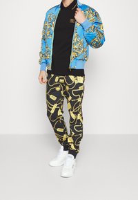 Man som bär en svart polo och svarta byxor med guldkedjemotiv, en blå och guldmönstrad jacka, samt vita sneakers.