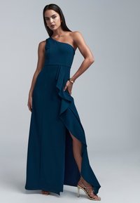 Ein schulterfreies, marineblaues Kleid aus strukturiertem Stoff, mit einem gerafften Vorderteil und einem hohen Schlitz bis zum Oberschenkel, kombiniert mit goldenen Riemchen-Heels.