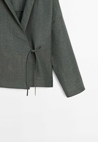 Blazer en lin gris avec un design croisé, fermeture par lien et manches trois-quarts. Tissu texturé avec une légère brillance et un détail de couture visible.